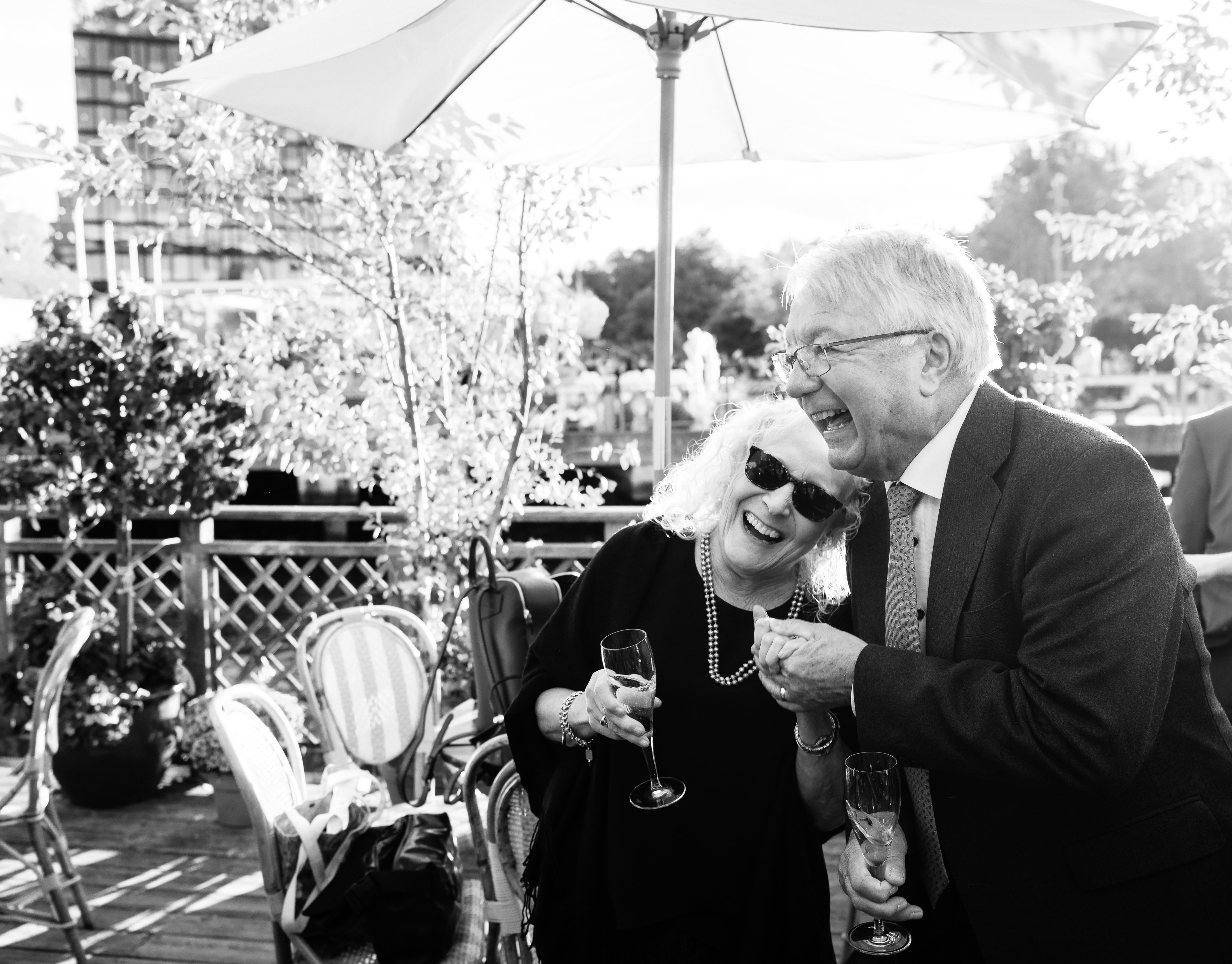 Black and white photograph of two guests laughing over champagne beneath a white umbrella at an outdoor party.