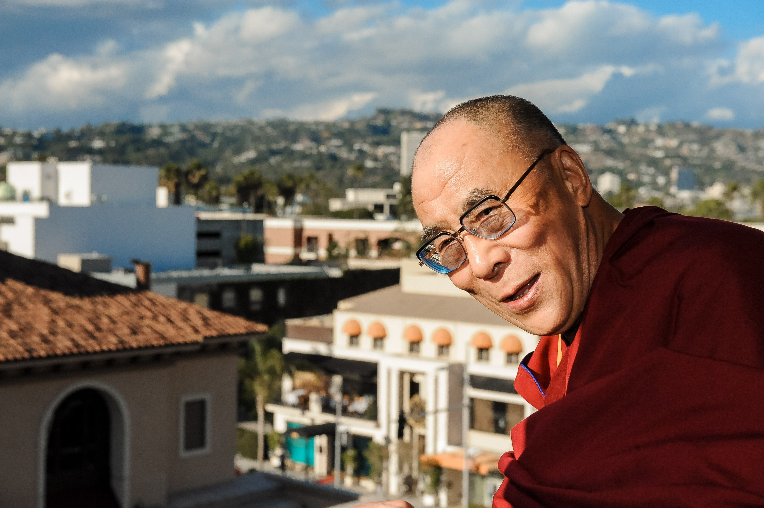 The Dalai Lama in maroon robes on a rooftop terrace, a hillside cityscape behind him.