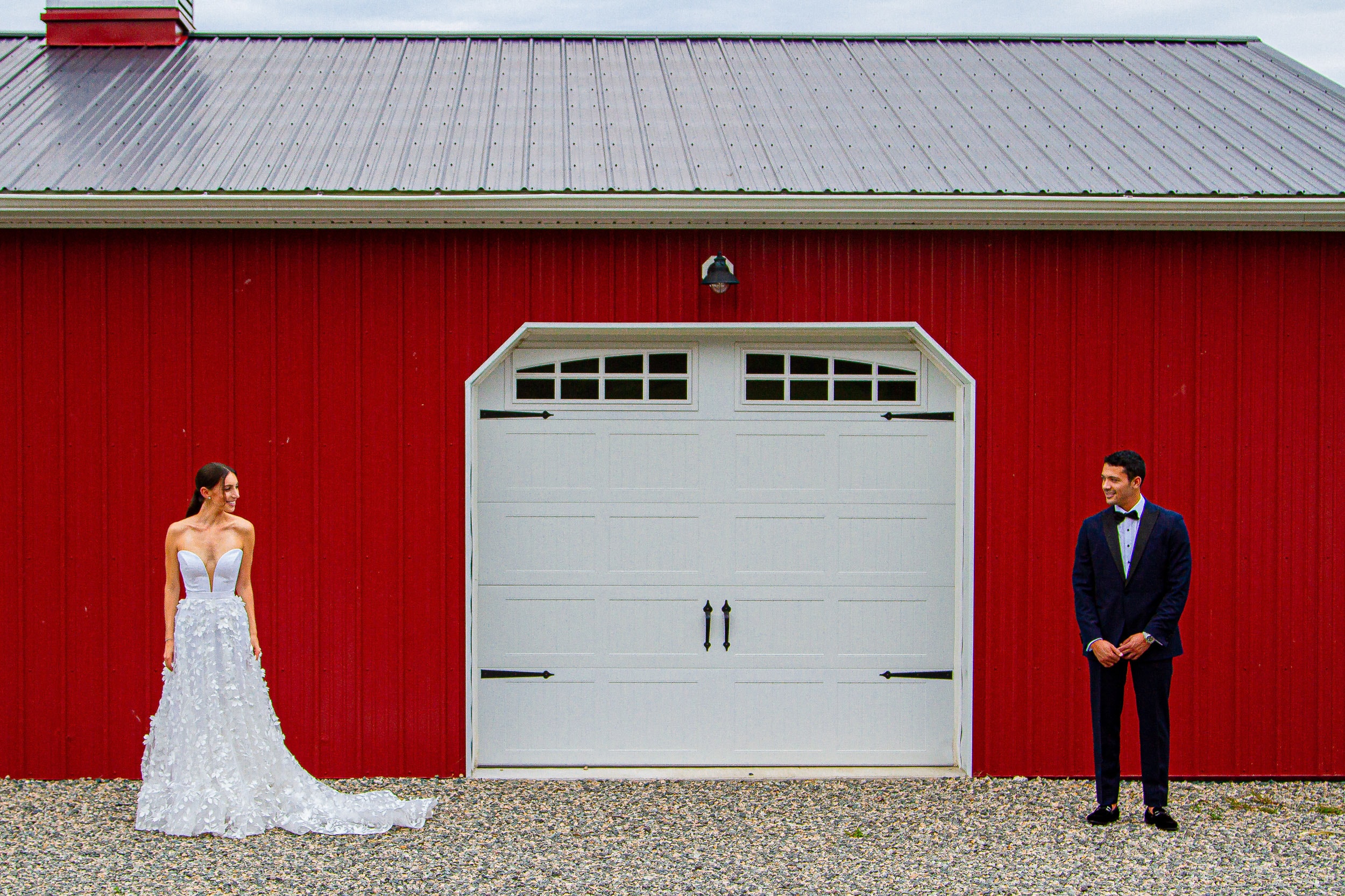 A bride and groom smiling at each other across the white door of a red barn before their first look.